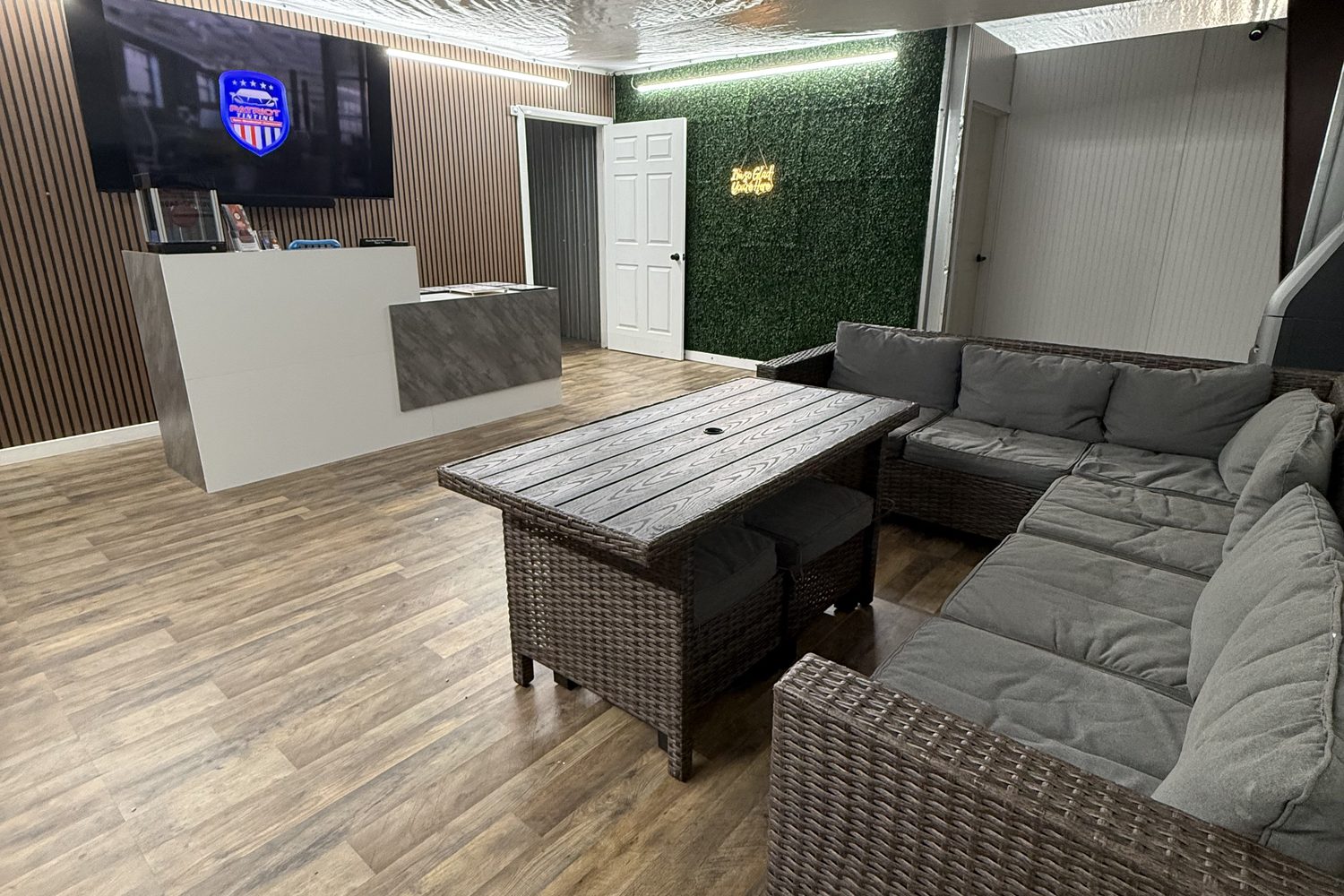The Patriot Tinting lounge in Hampton: L-shaped wicker sectional and coffee table, TV on the slat wall, front counter and a living green wall by the door